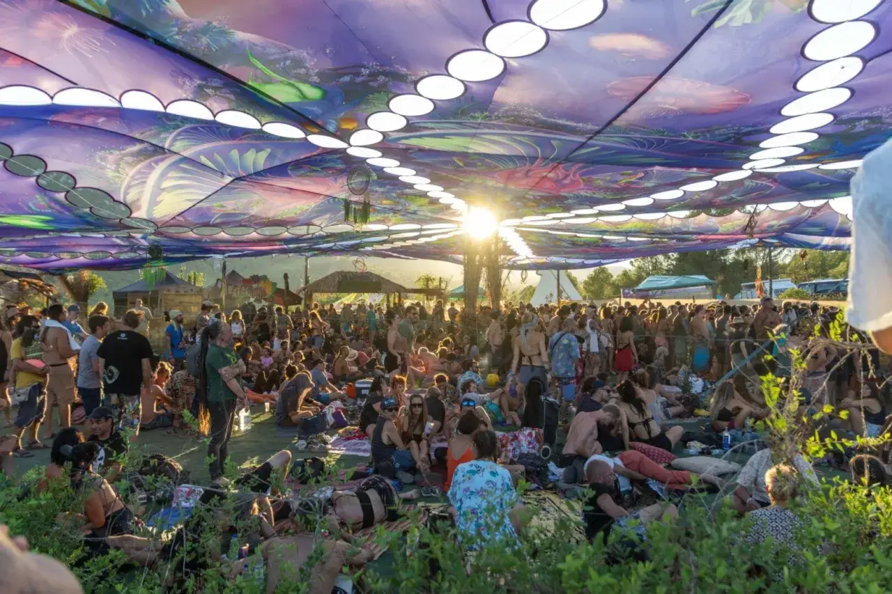 Multitud disfrutando bajo un toldo psicod&eacute;lico en un festival psytrance Espa&ntilde;a. El sol se filtra, creando un ambiente m&aacute;gico.