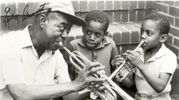 Louis Armstrong enseña a dos niños a tocar la trompeta. Ella Fitzgerald, aunque no está en la imagen, es una leyenda del jazz junto a él.