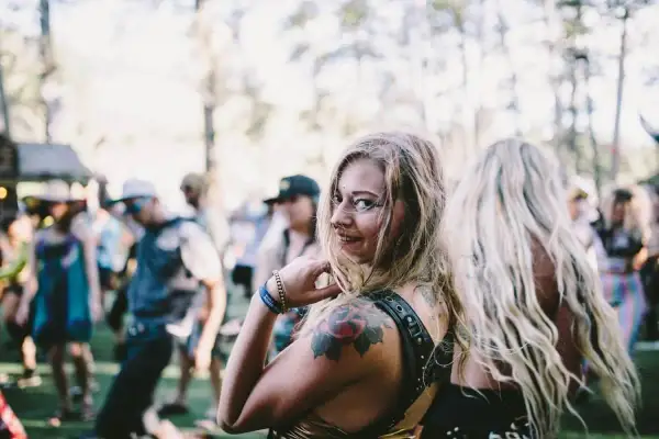 Chica sonriente con tatuajes en un festival de música electrónica, rodeada de gente bailando bajo el sol.