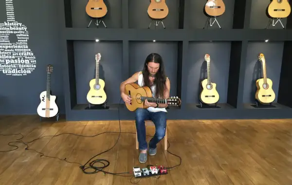 Un guitarrista con barba toca una guitarra acústica sentado frente a pedales de efectos.