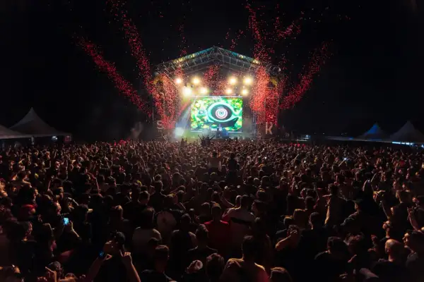 Multitud en un concierto al aire libre, con confeti rojo cayendo sobre el escenario y una pantalla gigante. El **bass** retumba en el parque.