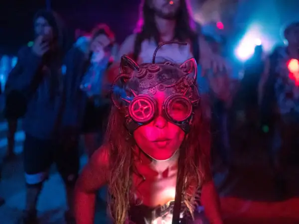 Una persona con una máscara de gato steampunk y gafas de aviador, rodeada de luces de neón y gente bailando. Así es que es la rave.