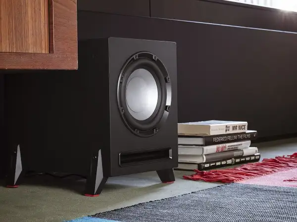 Subwoofer negro con detalles rojos, junto a libros y una alfombra.
