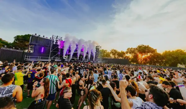 Multitud bailando en un festival al atardecer, con humo y luces moradas saliendo del escenario. ¡Un ambiente genial para los parkineos de donde es!