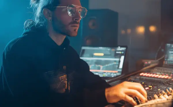 Hombre con gafas y coleta trabajando en un estudio de sonido, creando música electrónica para su blog.
