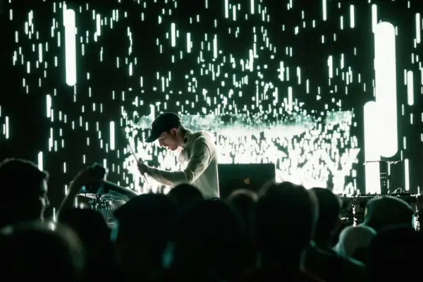 Un DJ techno en acción, con una pantalla de luces verdes y blancas parpadeantes detrás.