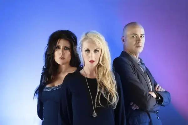 Miembros de The Human League: dos mujeres con maquillaje dramático y un hombre calvo con traje, posando ante un fondo azul y morado.
