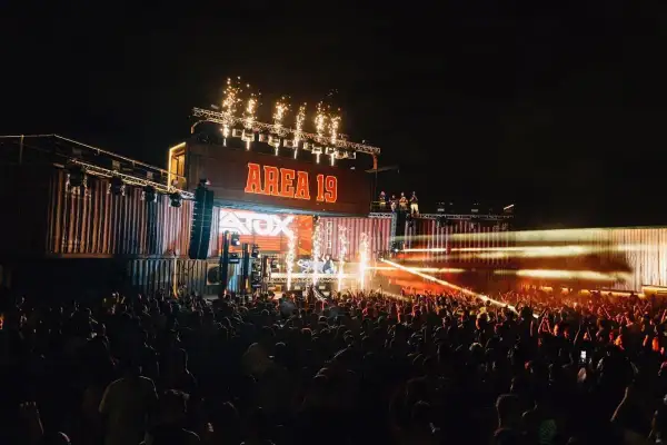 Una multitud celebra en un concierto nocturno, con fuegos artificiales y luces láser iluminando el escenario "AREA 19" y el **nexus fabrik** de contenedores.