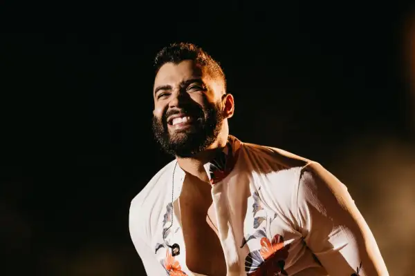 Un cantante brasileño famoso joven ríe con alegría en el escenario, con una camisa blanca estampada y un collar.